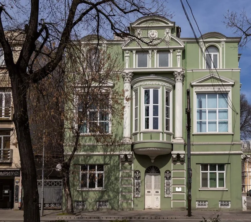 Фасада на зелена сграда с еркер, колони и декоративни фронтони.